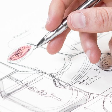 Close up of a hand holding silver pen over sketch of product packaging.